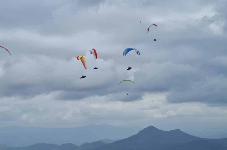 Zahlreiche bunte Gleitschirme beim Gruppenfliegen in einem Pulk vor einem bewölkten Himmel, was die Taktik des gemeinsamen Thermikfliegens für schnelle Streckenflüge illustriert.
