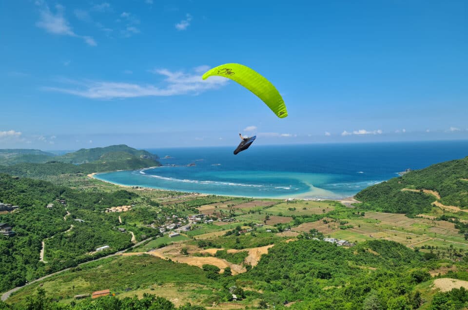 Ein Gleitschirmflieger mit einem neongrünen LittleCloud SuperFly 19,5 schwebt über der malerischen Küste von Lombok, Indonesien. Im Hintergrund sind eine Bucht mit türkisfarbenem Wasser und üppig grüne Hügel unter einem blauen Himmel zu sehen.