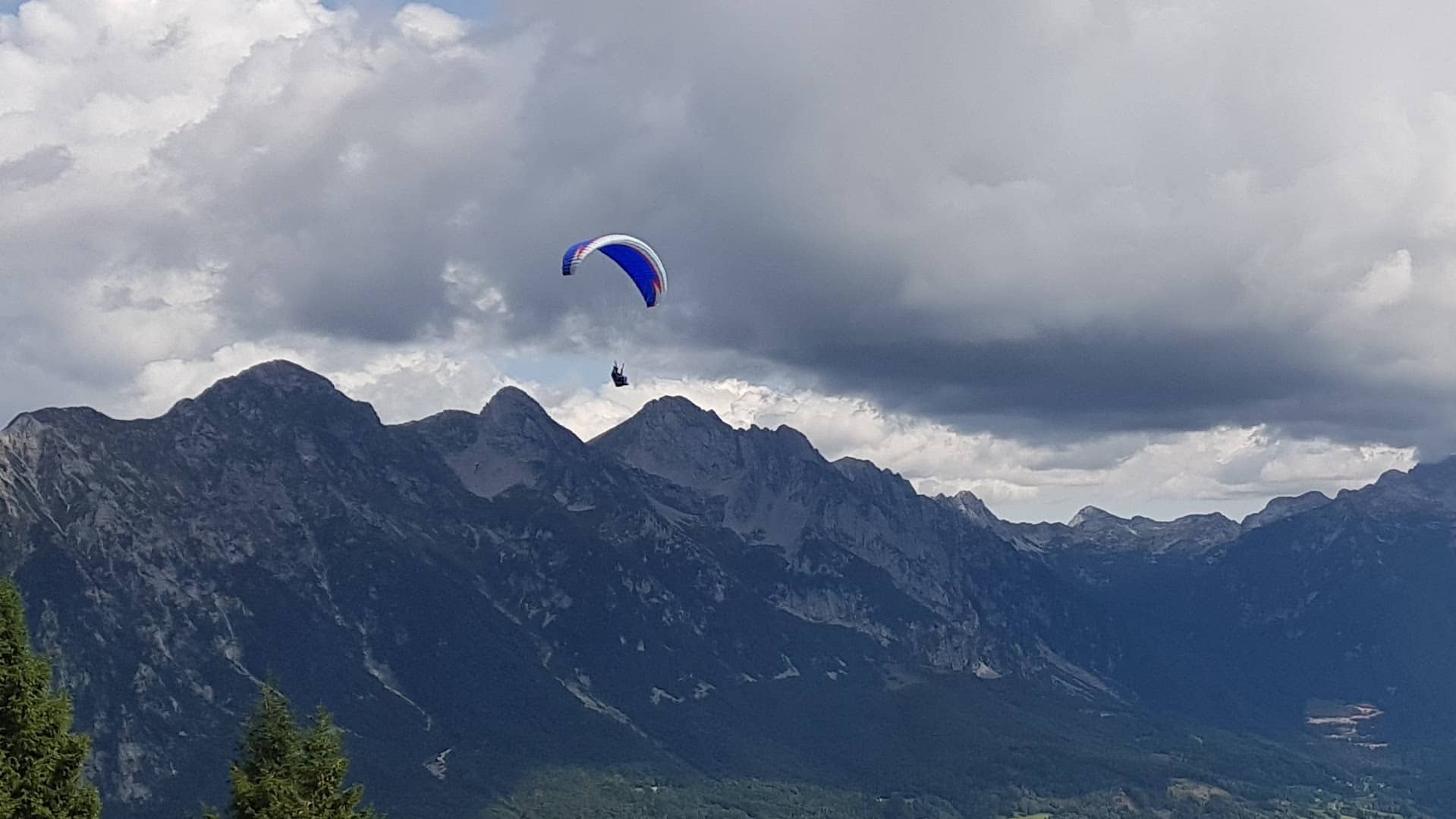 Ein einzelner Gleitschirmflieger mit einem blau-weißen Low-B-Schirm fliegt sicher vor einer imposanten, dunklen Bergkette unter einem dramatischen Wolkenhimmel, der einen thermisch aktiven Tag andeutet.
